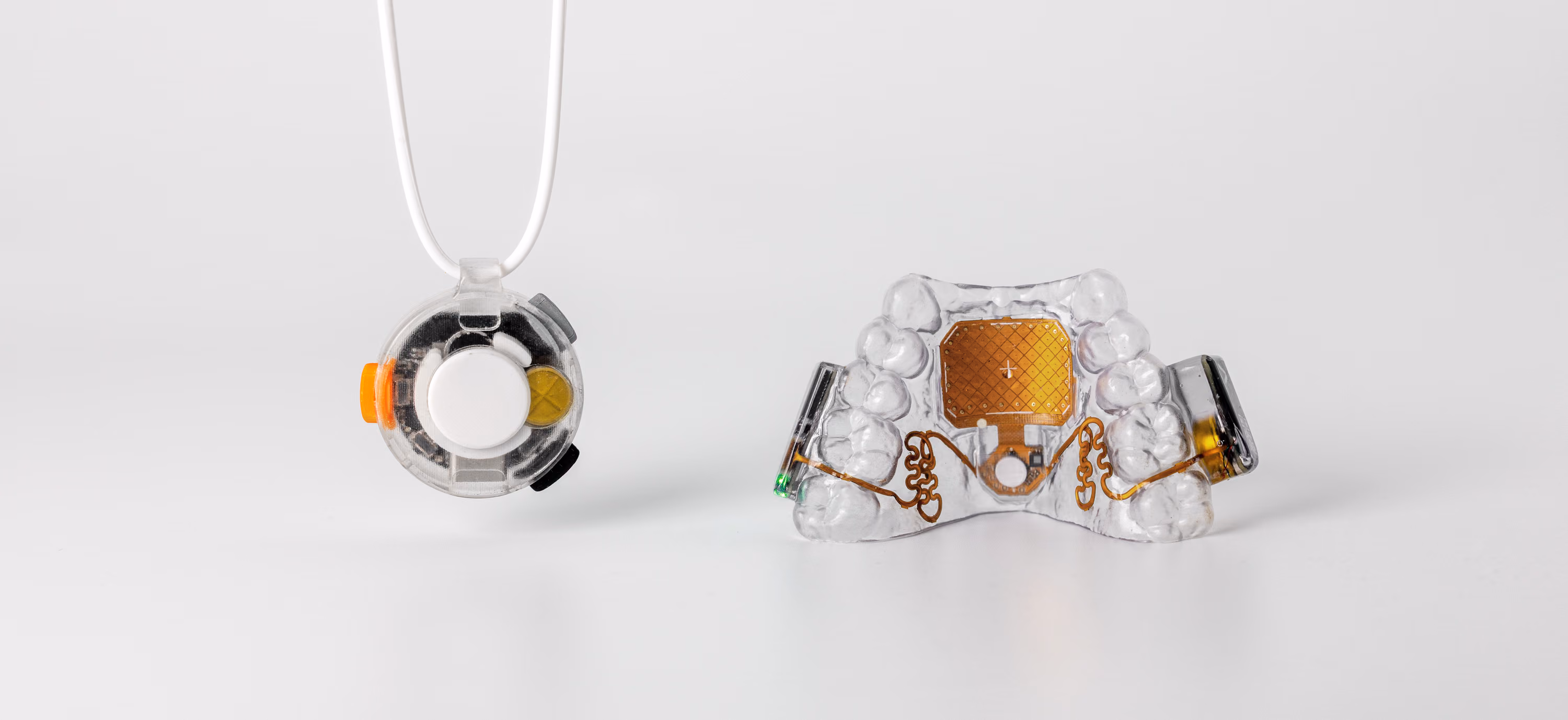 VOX and the MouthPad side by side on a pale studio background. VOX hangs at the left on its cord: a small round device in a clear housing, its orange side control and pale capture button visible through the case. The MouthPad sits at the right: a clear custom-fit retainer with a gold touchpad at its centre and gold circuitry curving out to the side electronics.