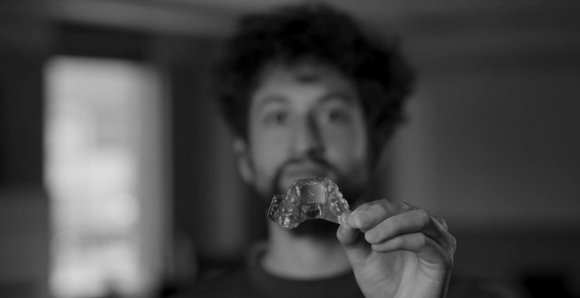 Tomás looks up with his mouth open, the MouthPad fitted to the roof of his mouth. In his other hand a phone shows the MouthPad app, its touchpad and force readout moving as his tongue drives the cursor.